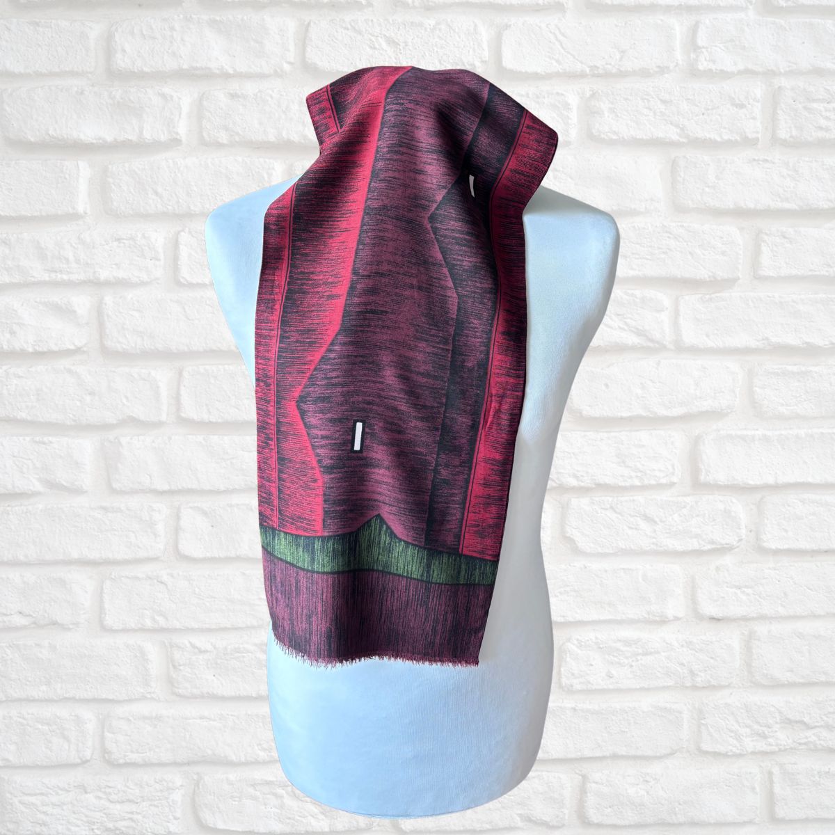 A patterned knit scarf in shades of red , dark purple, and green, draped on a light blue mannequin torso against a white brick wall background.