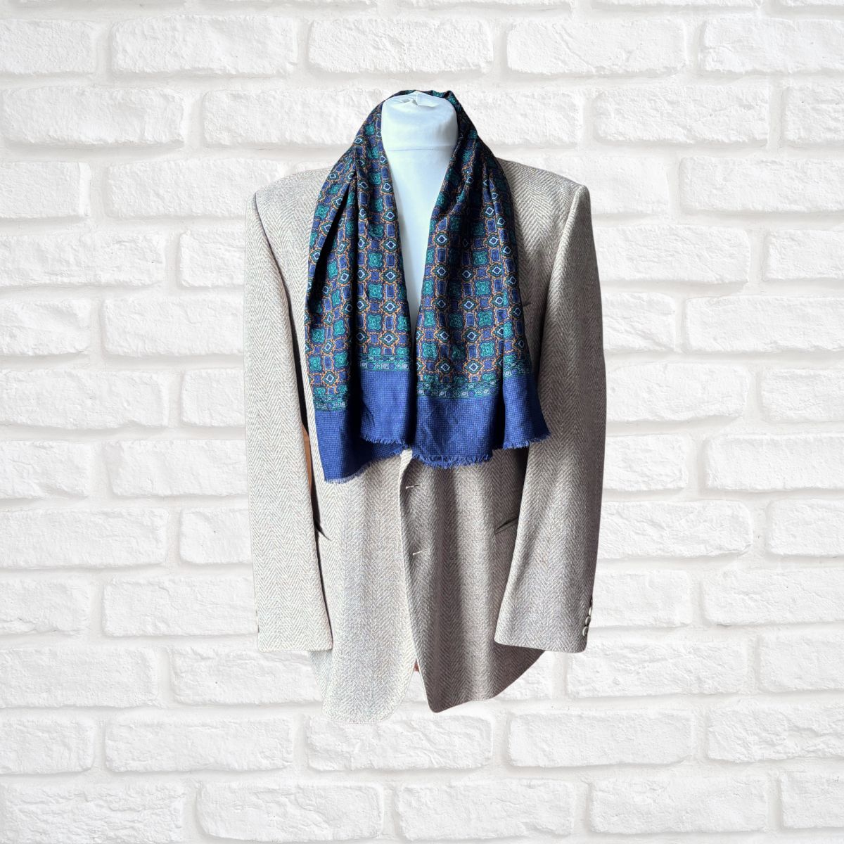 A patterned blue scarf with geometric designs in red, green, and yellow, draped around a light gray blazer on a white mannequin torso against a white brick wall background.