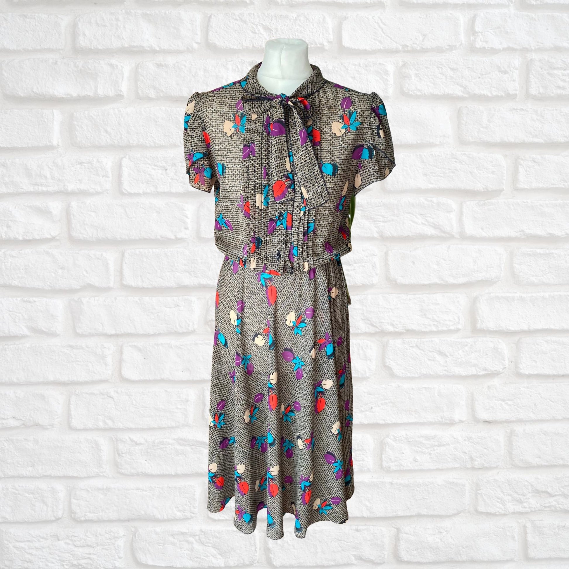 A vintage botanical print dress with a pleated bodice, black buttons, and a pussy bow collar, displayed on a mannequin against a white brick wall background.