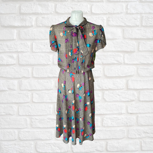 A vintage botanical print dress with a pleated bodice, black buttons, and a pussy bow collar, displayed on a mannequin against a white brick wall background.