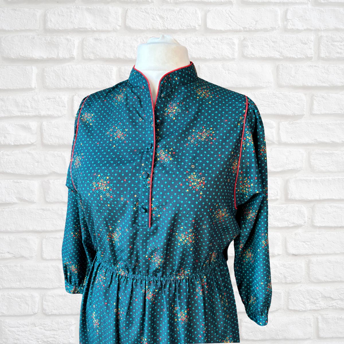 Teal dress with geometric pattern on a mannequin against a white brick wall.