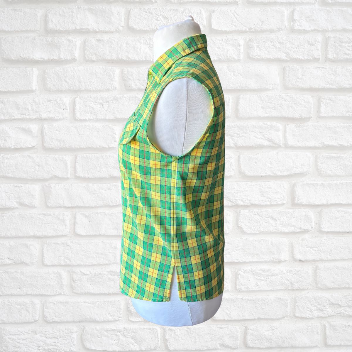 Green and yellow plaid sleeveless shirt on a mannequin against a white brick wall.