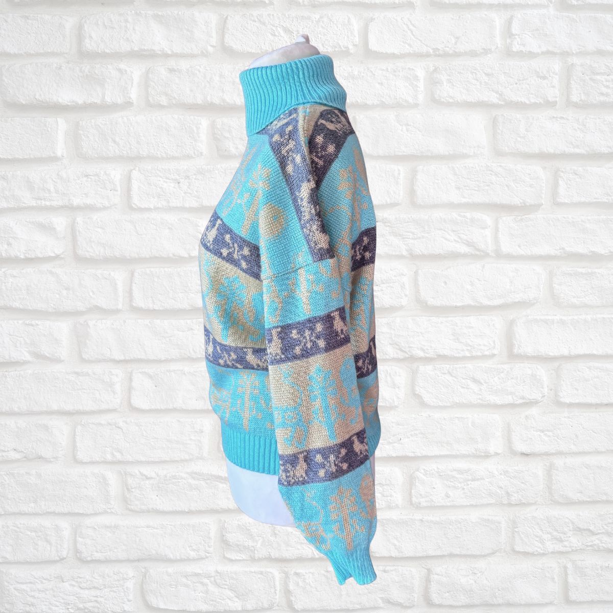 A turquoise blue turtleneck sweater with a beige and dark blue Nordic pattern, displayed on a white mannequin torso against a white brick wall background.