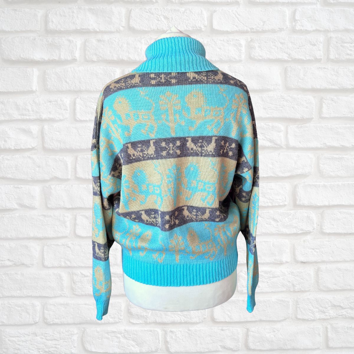 A turquoise blue and beige knit sweater with a dark gray patterned band, displayed on a white mannequin torso against a white brick wall background.