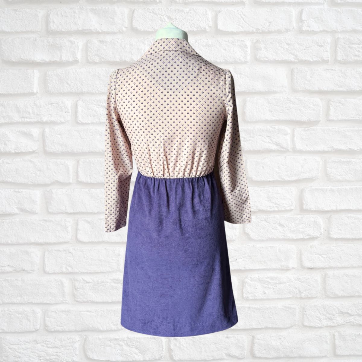 Mannequin wearing a two-tone dress with a geometric print top and solid purple skirt against a white brick wall.
