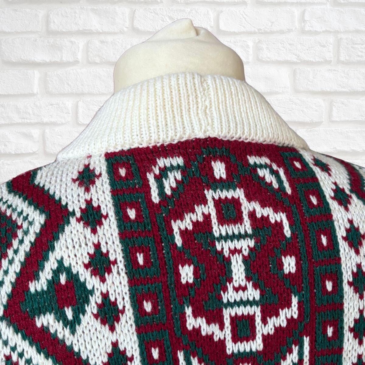 A close-up of a knit sweater on a cream-colored mannequin torso, showing a festive Christmas pattern in burgundy red, white, and dark green. The background is a white brick wall.