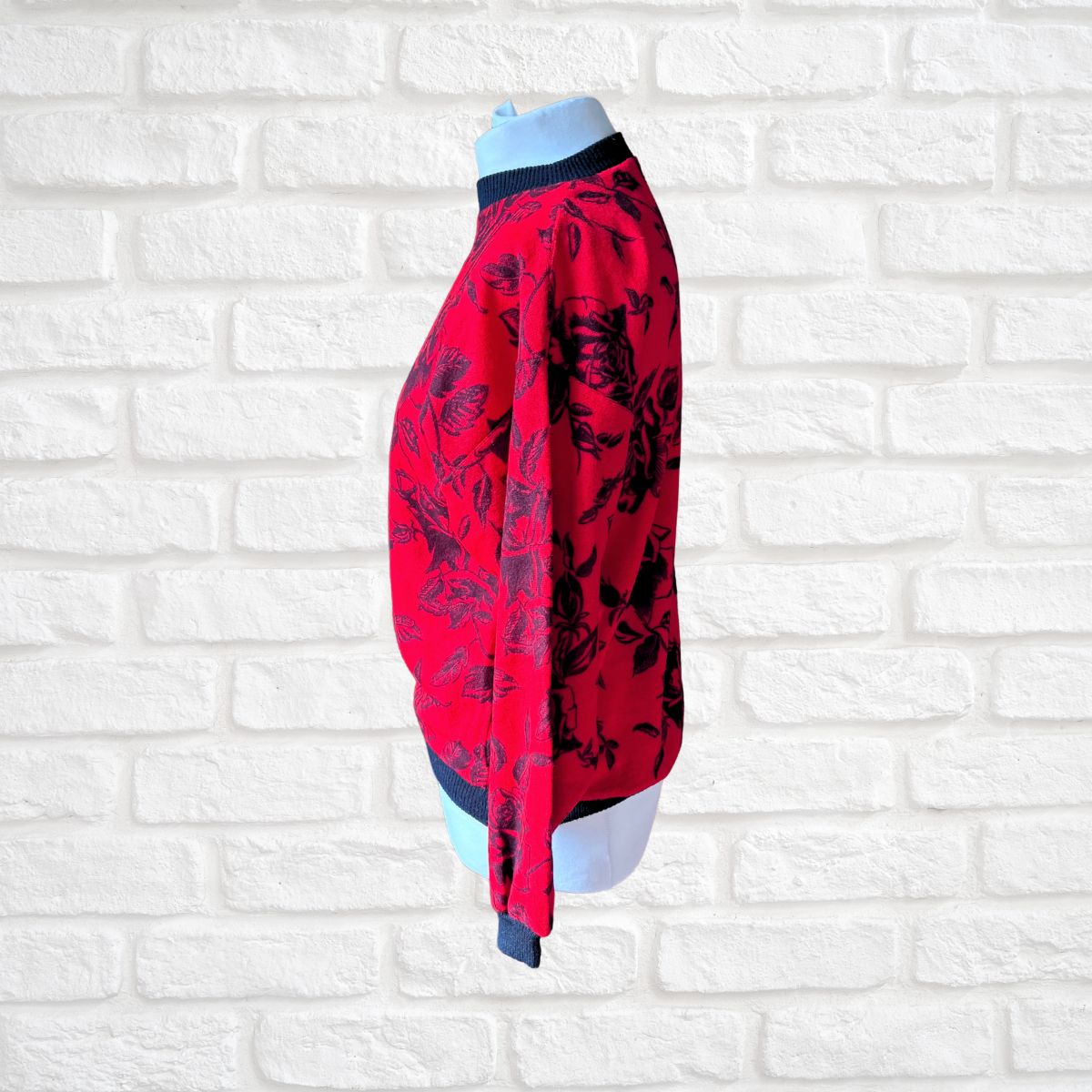 A red long-sleeve blouse with a black floral pattern, displayed on a light blue mannequin torso against a white brick wall background.