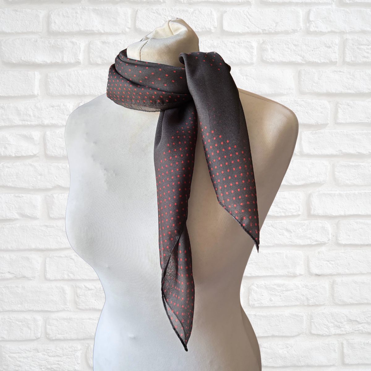 Mannequin wearing a dark gray scarf with red polka dots against a white brick wall.