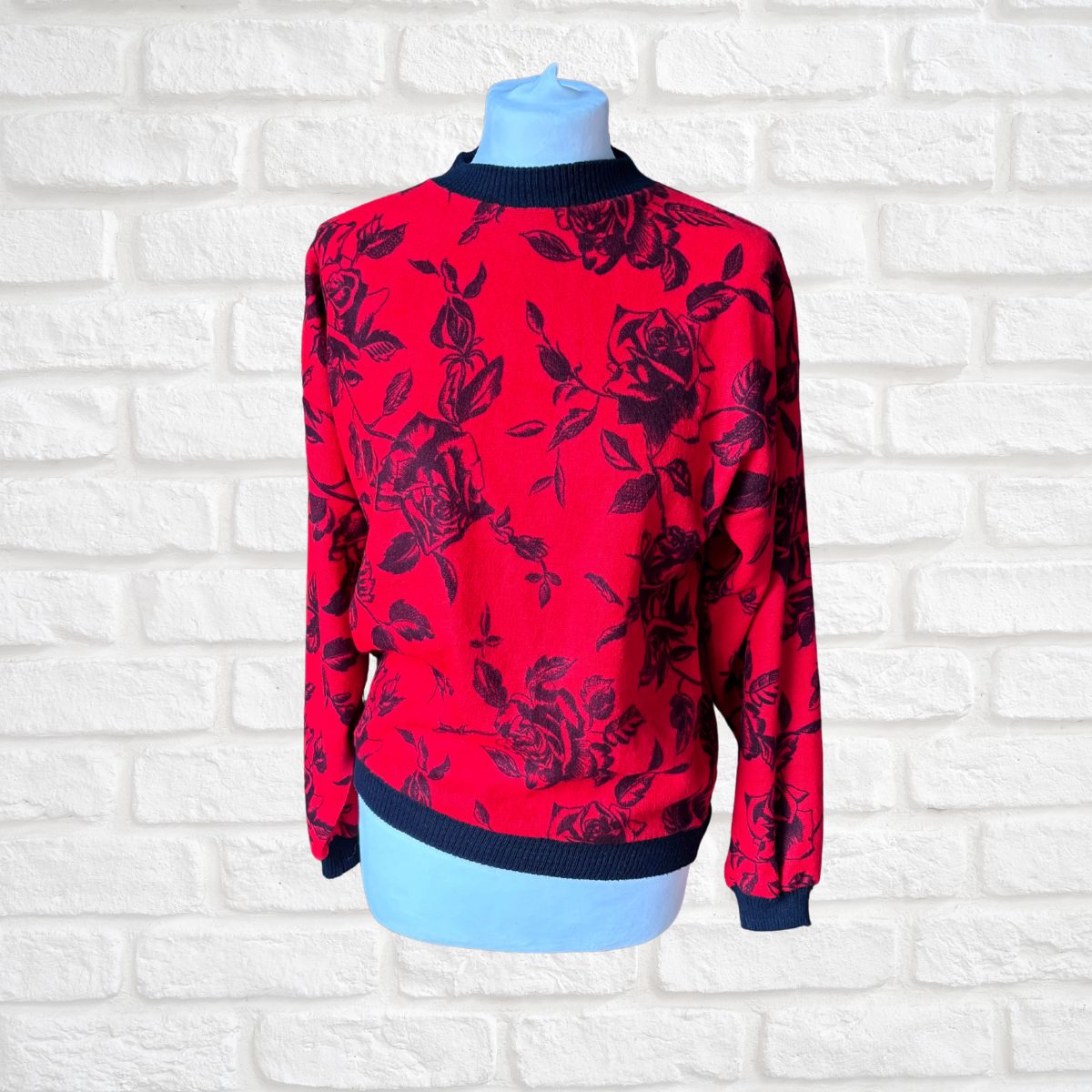 A red long-sleeve knit sweater with a black rose floral pattern, displayed on a light blue mannequin torso against a white brick wall background.