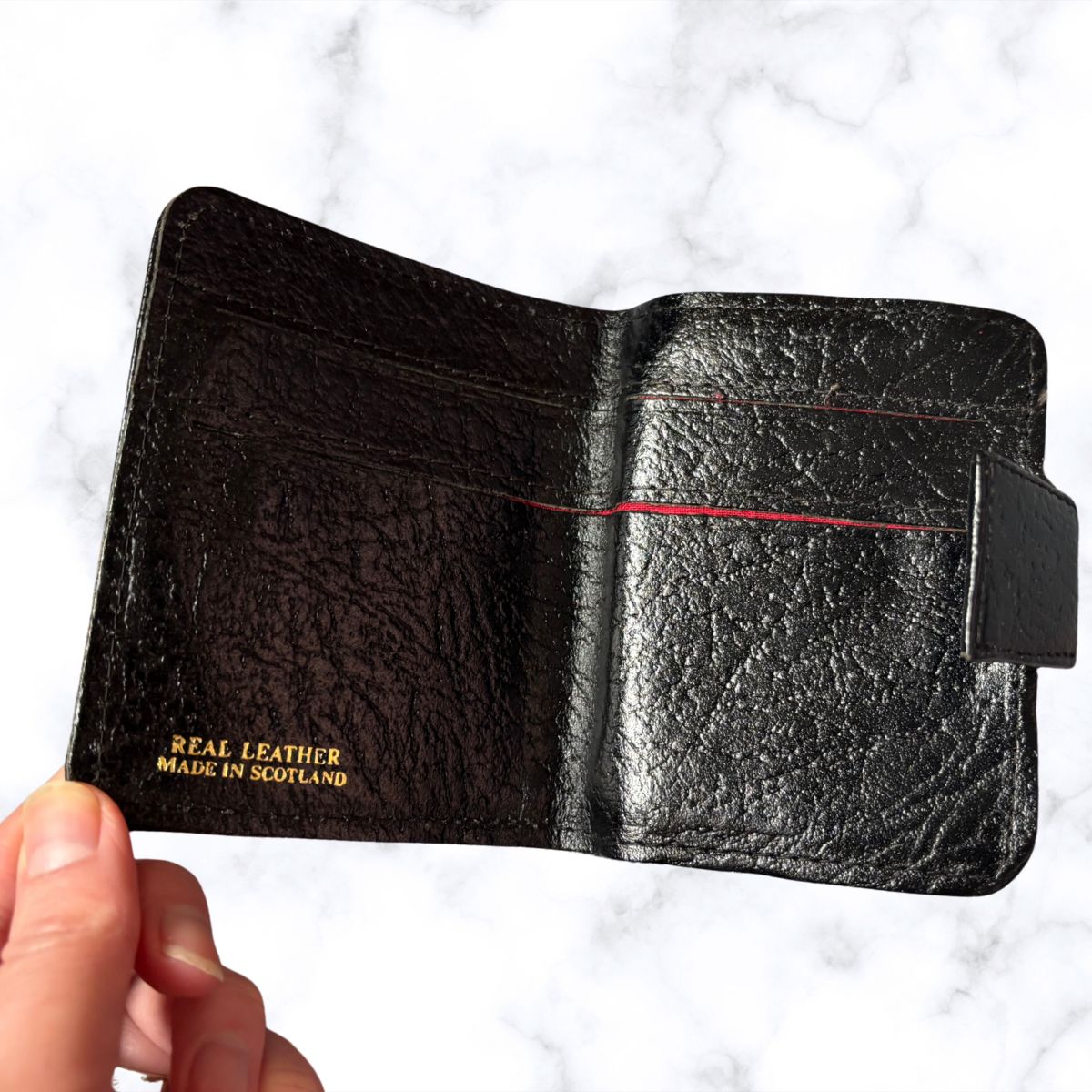 Black leather wallet held by a hand with 'Real Leather Made in Scotland' text on a white marble background.
