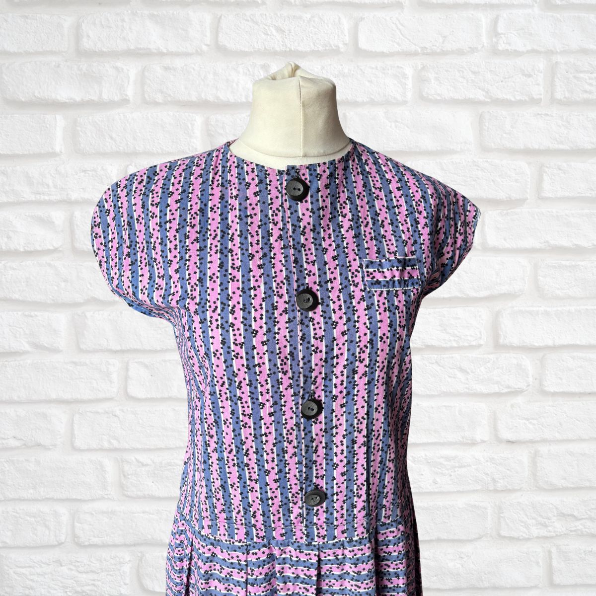 Purple and pink patterned dress on a mannequin against a white brick wall.