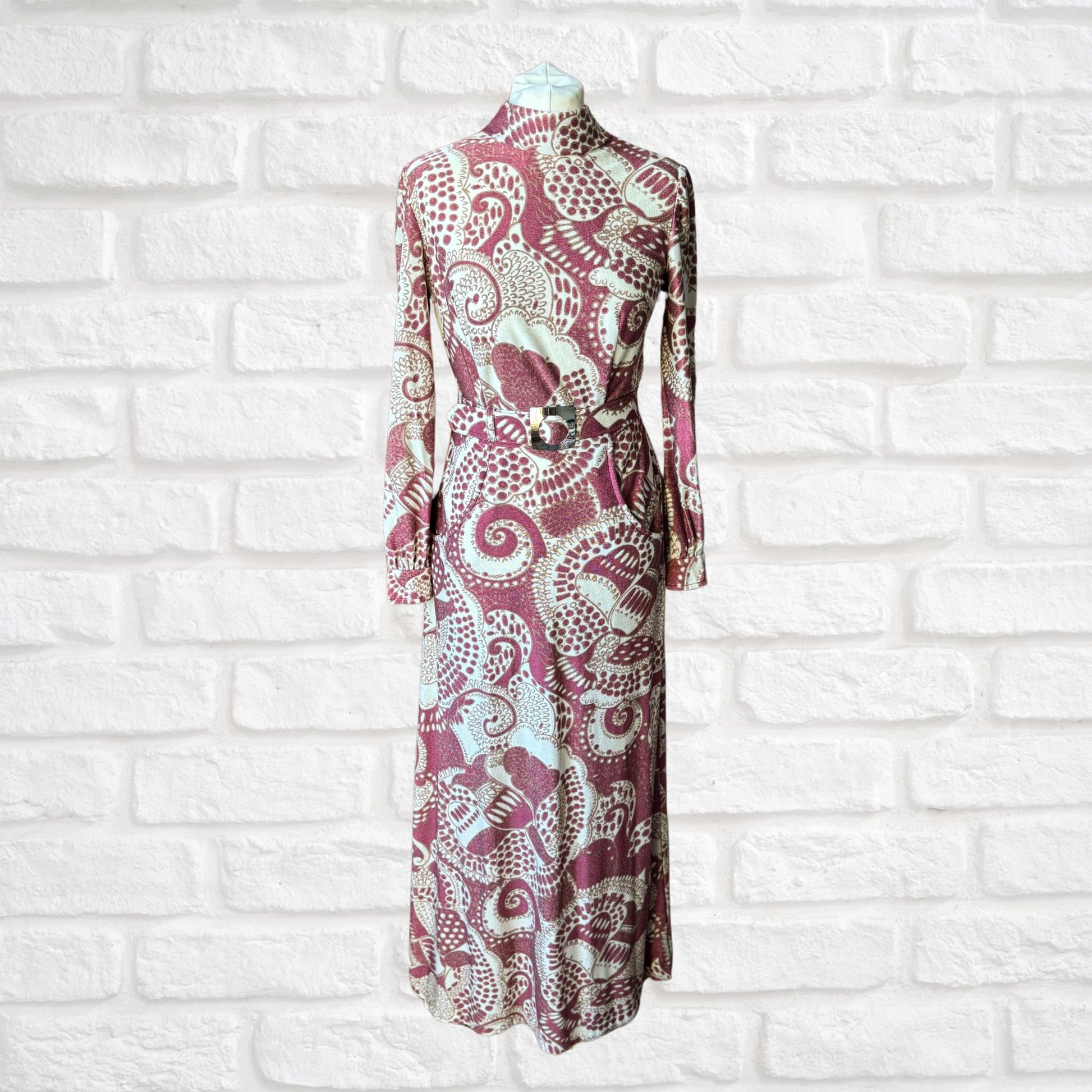 A vintage 70s long-sleeved maxi dress in a cream, burgundy, and brown abstract print, with a high turtle neck, long sleeves with silver button fastened cuffs, and a matching belt, displayed on a mannequin against a white brick wall background.