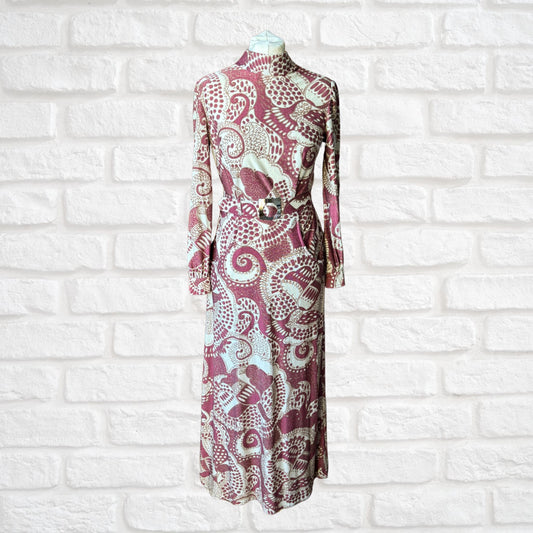 A vintage 70s long-sleeved maxi dress in a cream, burgundy, and brown abstract print, with a high turtle neck, long sleeves with silver button fastened cuffs, and a matching belt, displayed on a mannequin against a white brick wall background.