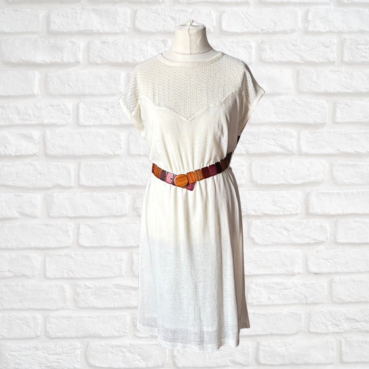 Cream dress with a brown  belt on a mannequin against a white brick wall.
