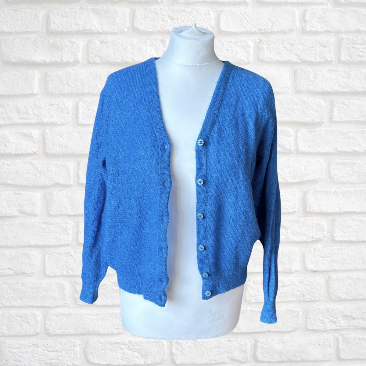A bright blue textured knit cardigan sweater displayed on a white mannequin torso against a white brick wall background.