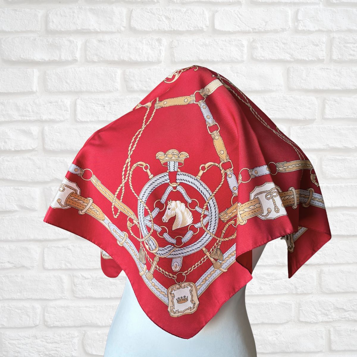 Red scarf with gold and white patterns on a mannequin against a white brick wall.
