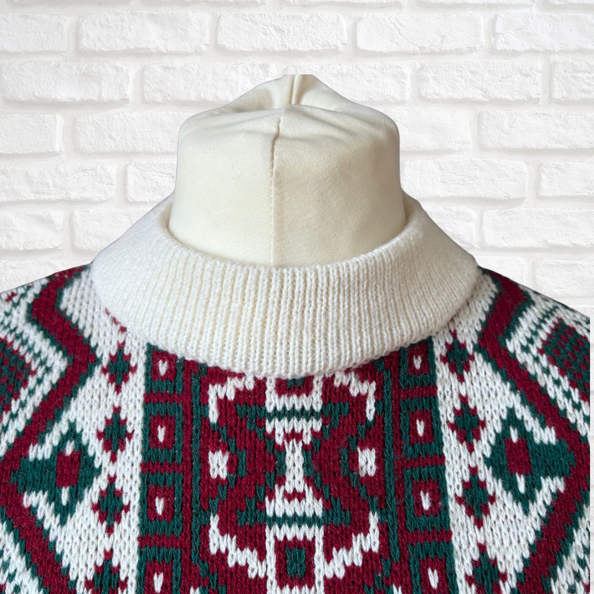 A close-up of a knit sweater on a cream-colored dress form, featuring a red, green, and white geometric pattern with a cream ribbed turtleneck, set against a white brick wall background.