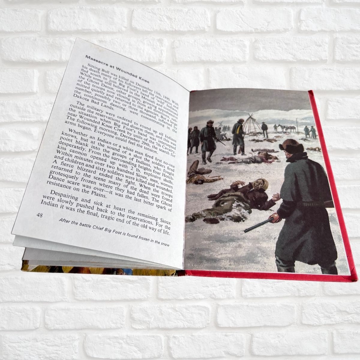 An open book showing printed text on the left page and a historical illustration on the right page, photographed against a white brick wall background.