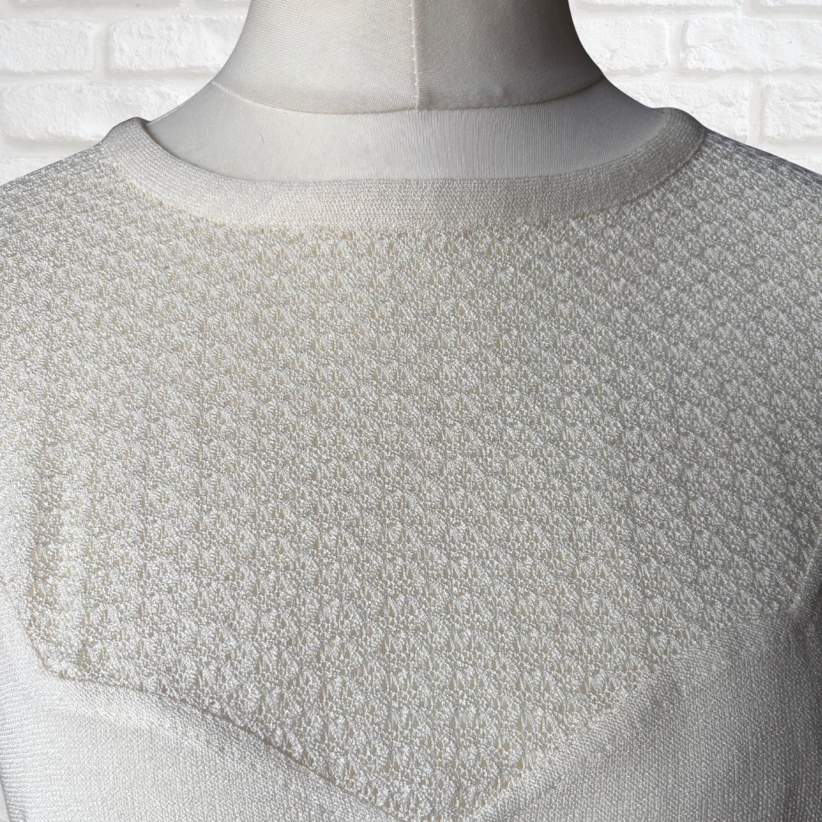 Textured white garment on a mannequin against a white brick wall.