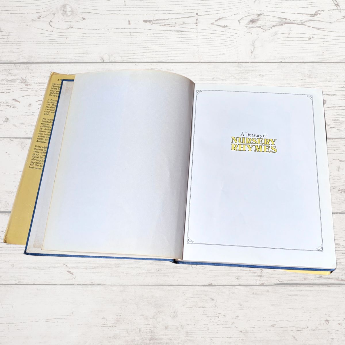 An open hardcover book titled 'A Treasury of Nursery Rhymes' lies on a light whitewashed wooden surface. The left page is blank, while the right page displays the gold title.