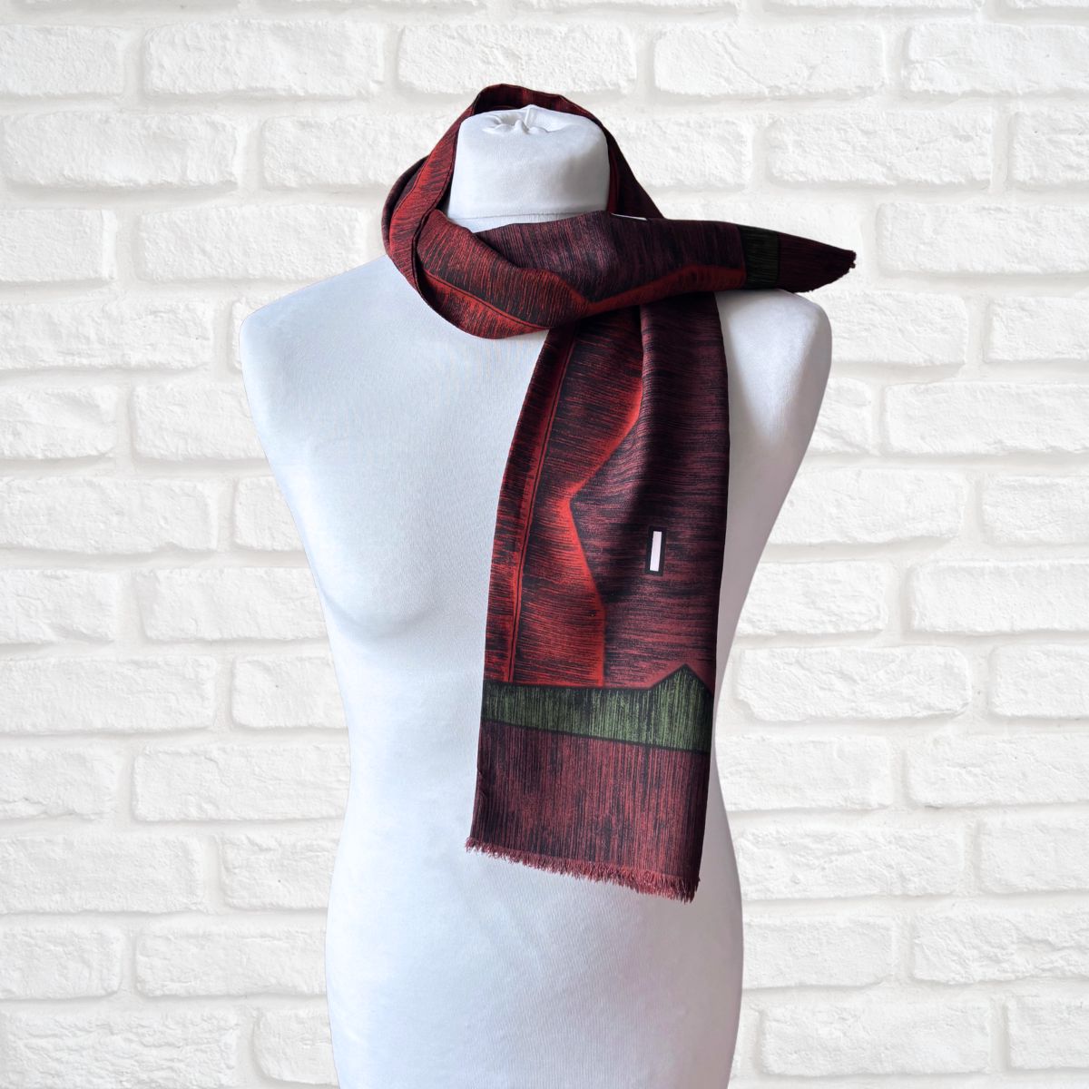 A patterned scarf in deep red and black with a green landscape-like band near the hem, draped on a white mannequin torso against a white brick wall.