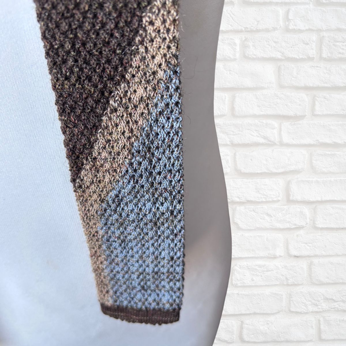 Knitted tie with gradient color pattern on a mannequin against a white brick wall.