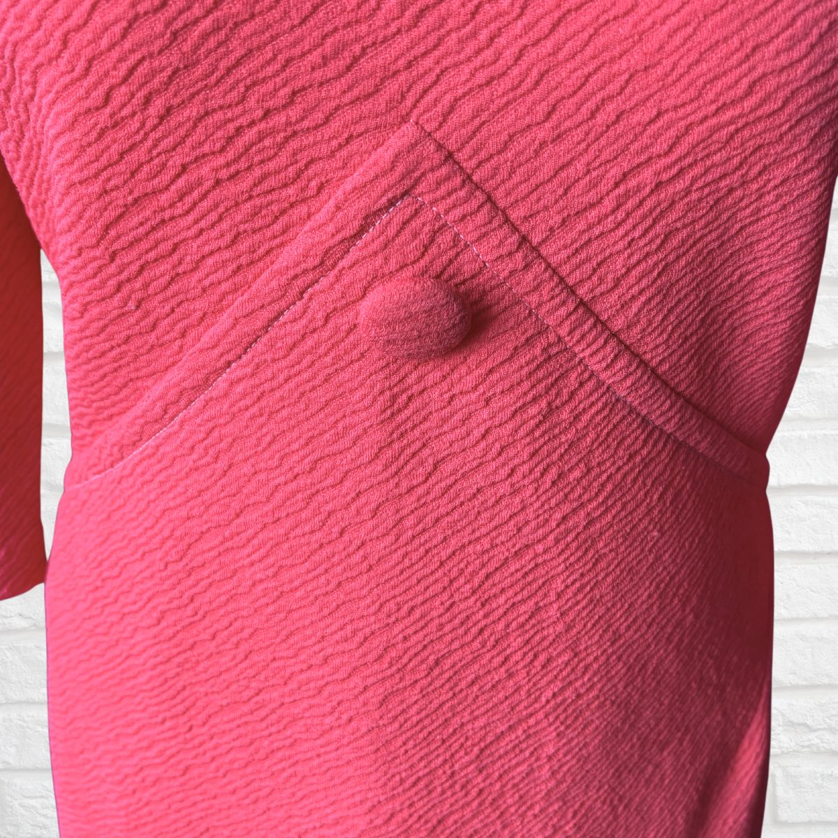 A close-up of a textured pink dress featuring a matching fabric-covered button, set against a white brick wall background.