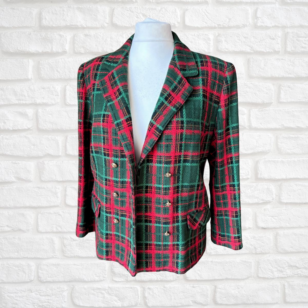A double-breasted plaid blazer in green, red, and black tartan, displayed on a white mannequin torso against a white brick wall background.