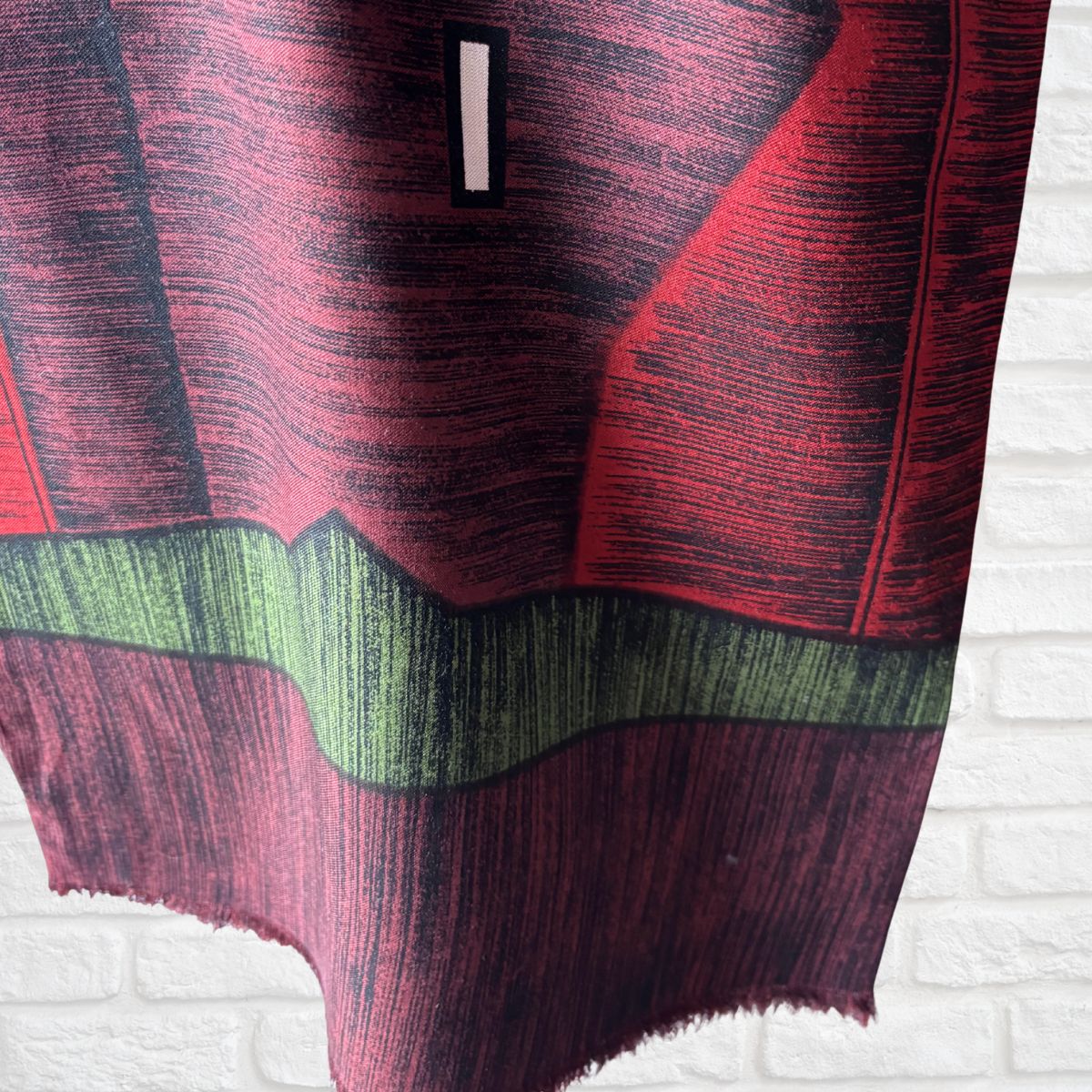 A close-up of a scarf with a red and black textured design, a horizontal green band, and a small black rectangular accent, shown against a white brick wall background.