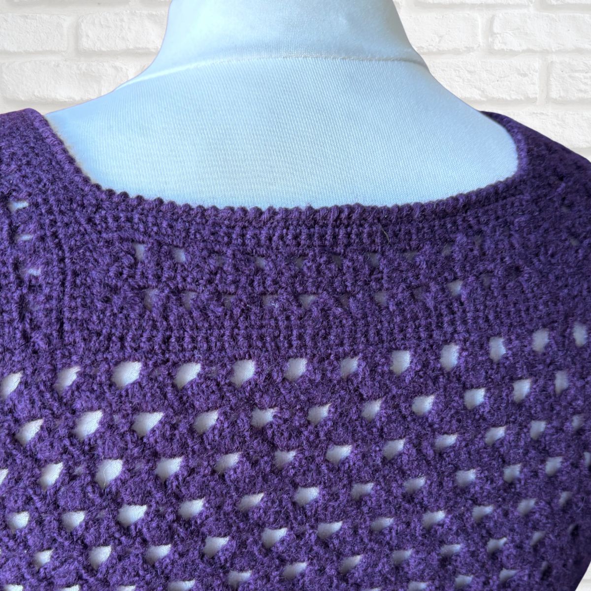 Close-up of a purple crochet top on a mannequin with a white brick wall background