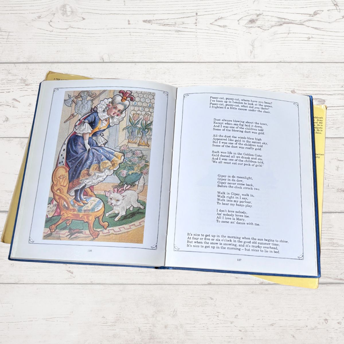 An open vintage children's book lying flat on a white wooden surface, showing a colorful illustration of a girl in a blue dress on the left page and printed text on the right page.