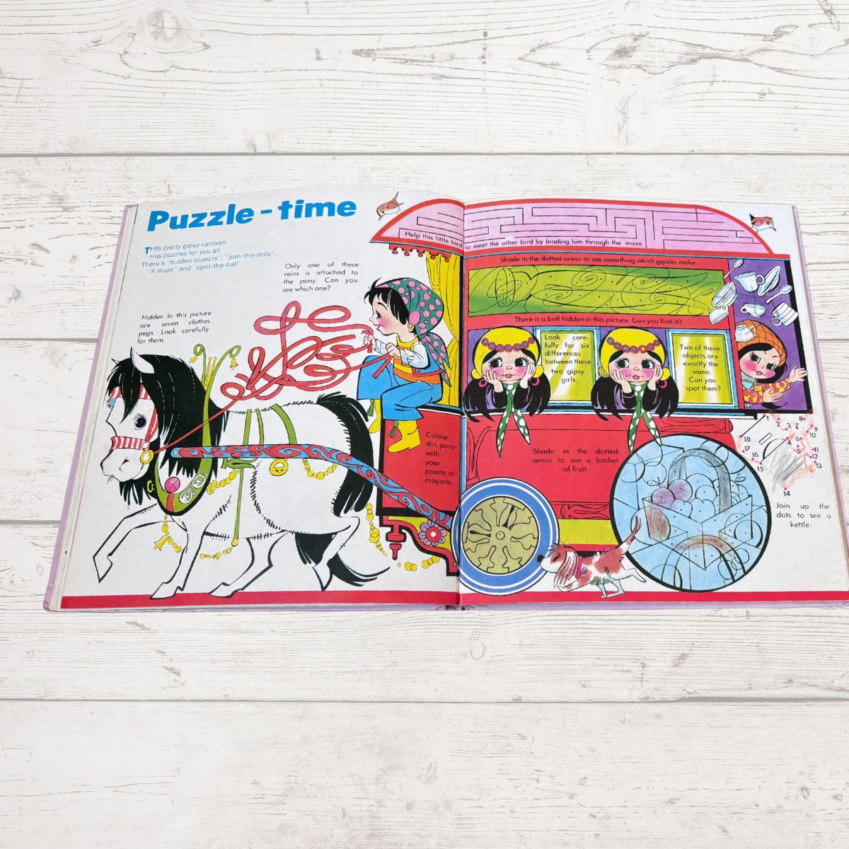 An open children's puzzle book titled 'Puzzle-time' laid flat on a white wood surface. The spread shows colorful cartoon illustrations, including a horse, a pink carriage, and several children, with text and puzzle elements visible on both pages.