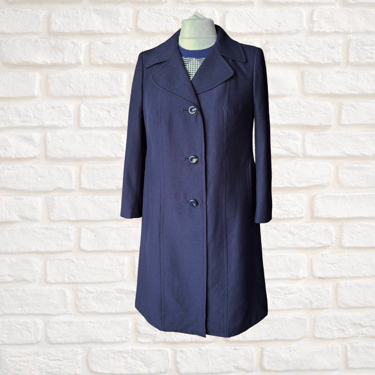 A navy blue knee-length coat with notched lapels and three front buttons, displayed on a mannequin torso against a white brick wall background.