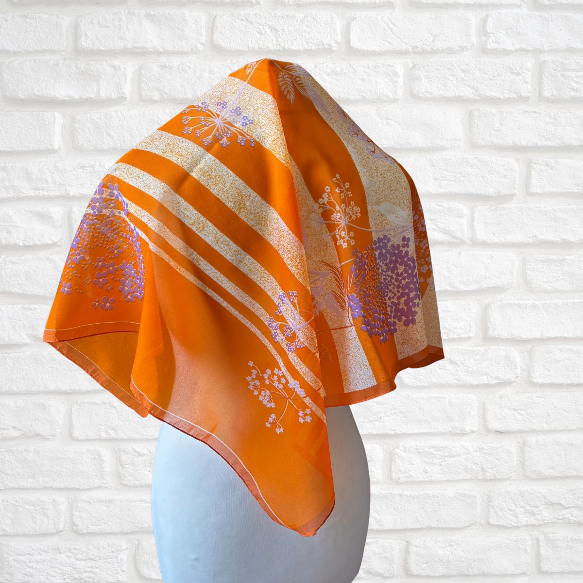 Orange scarf with floral patterns draped over a mannequin head against a white brick wall.