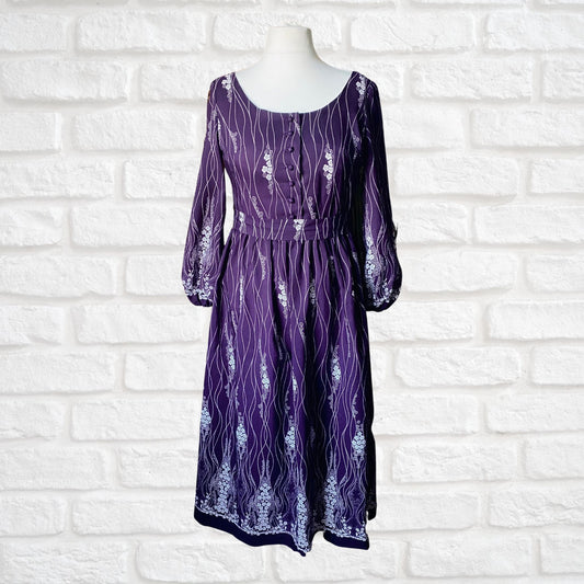 A purple 70s vintage maxi dress with white floral print, featuring a scooped neckline, button down bodice, and balloon sleeves, displayed on a mannequin against a white brick wall background.