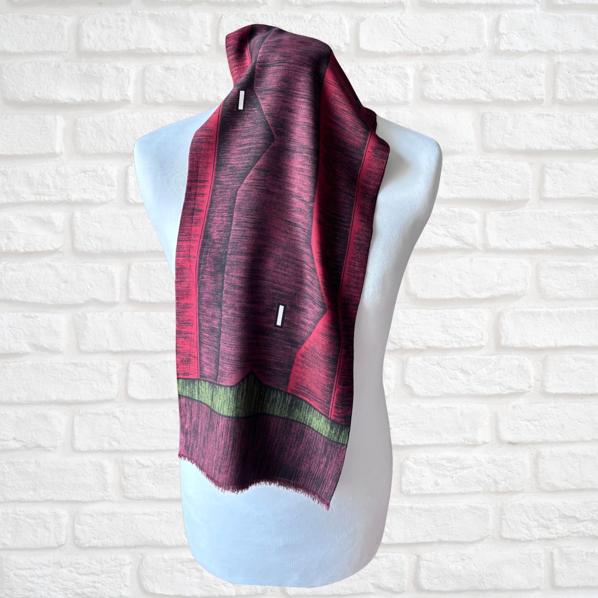 A patterned rayon scarf draped on a white mannequin torso, set against a white brick wall background. The scarf features a geometric design in deep burgundy, dark purple, and a horizontal olive green stripe near the bottom.