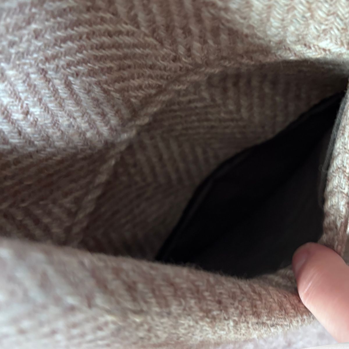 Close-up of a beige and taupe herringbone woven jacket , showing the texture and interior pocket opening with a finger pointing inside.