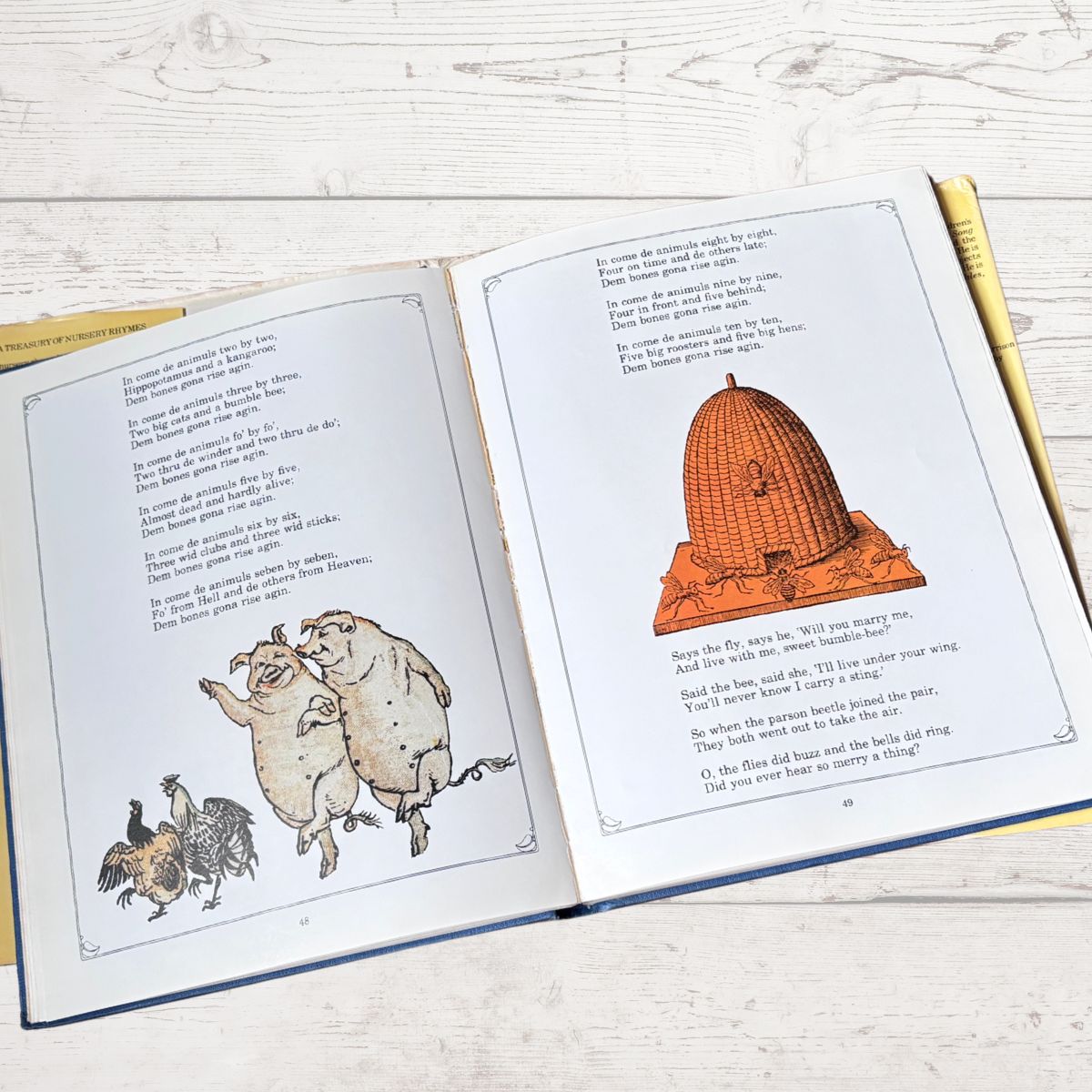 An open children's book lying on a light wood surface, showing printed text and illustrations including pigs, chickens, and a beehive.