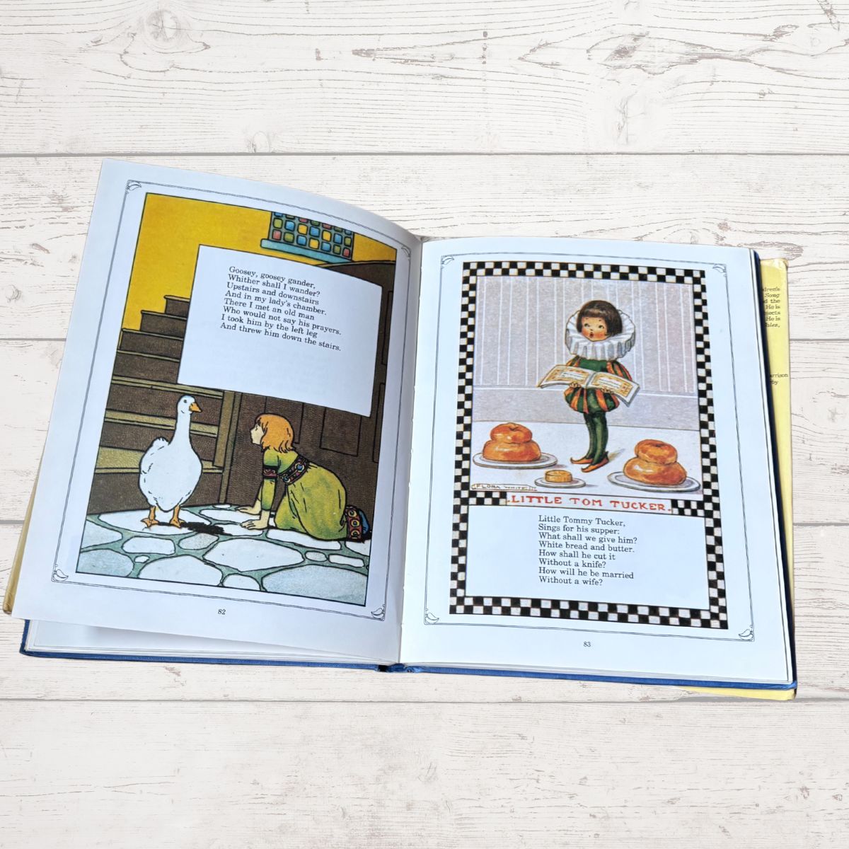 An open children's book lying on a light wood surface, showing two illustrated pages with text boxes and cartoon drawings. The left page depicts a white goose and a small dog, and the right page shows a child character with food