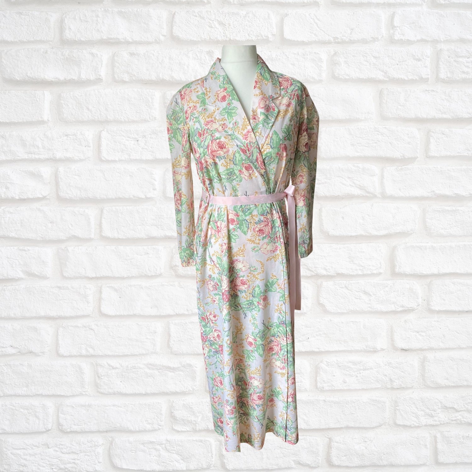 A vintage 80s full-length pink rose print dressing gown with a pink ribbon belt, displayed on a mannequin against a white brick wall.