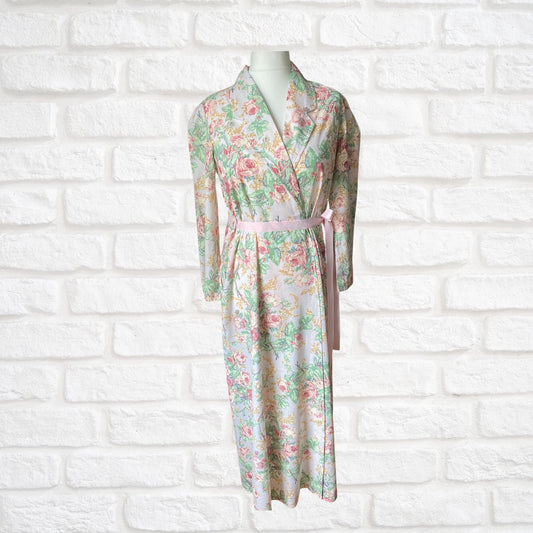A vintage 80s full-length pink rose print dressing gown with a pink ribbon belt, displayed on a mannequin against a white brick wall.