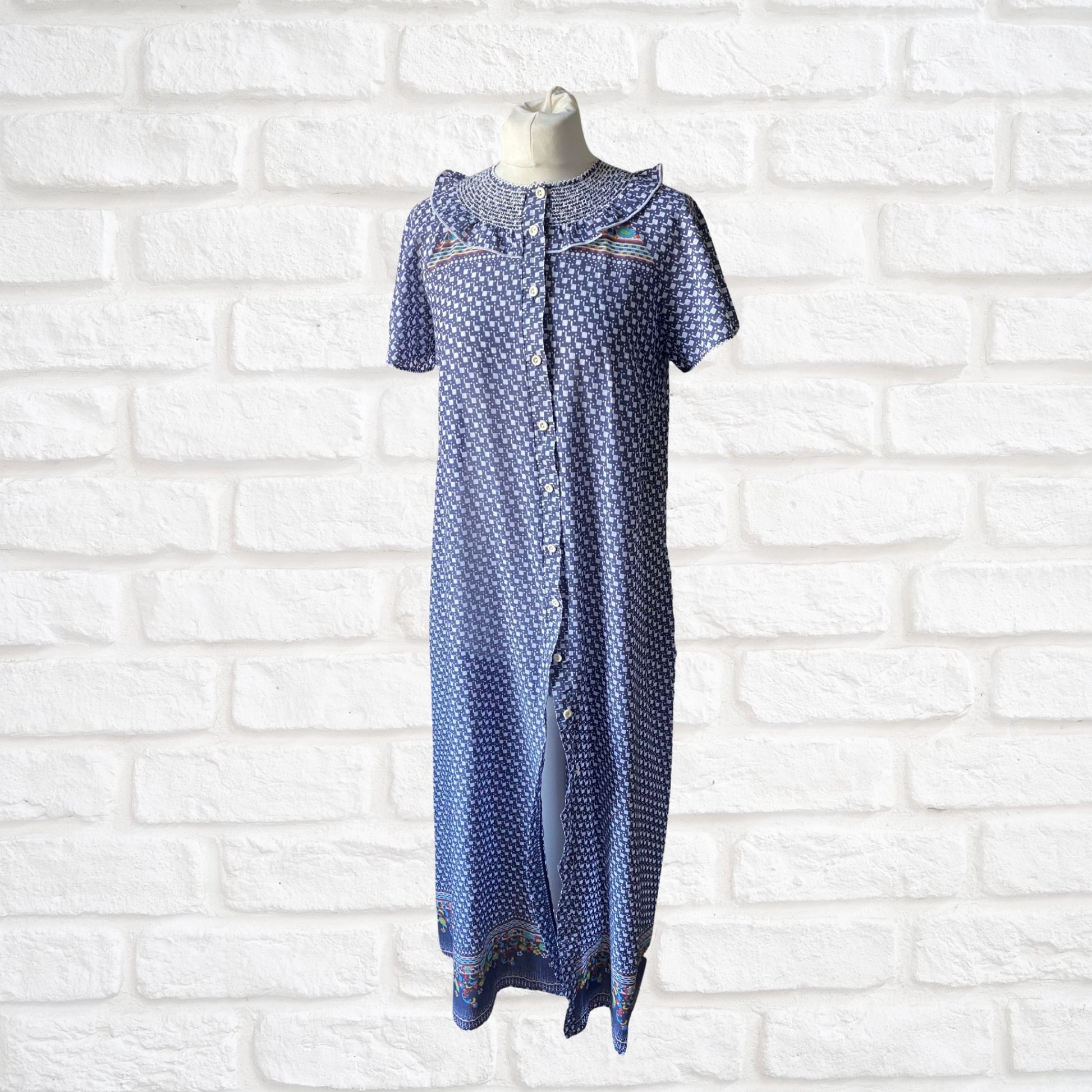 A vintage 70s blue and white geometric print dressing gown with multicolored florals, featuring short sleeves, bodice smocking, and a frill detail, displayed on a mannequin against a white brick wall.