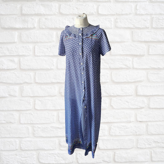 A vintage 70s blue and white geometric print dressing gown with multicolored florals, featuring short sleeves, bodice smocking, and a frill detail, displayed on a mannequin against a white brick wall.