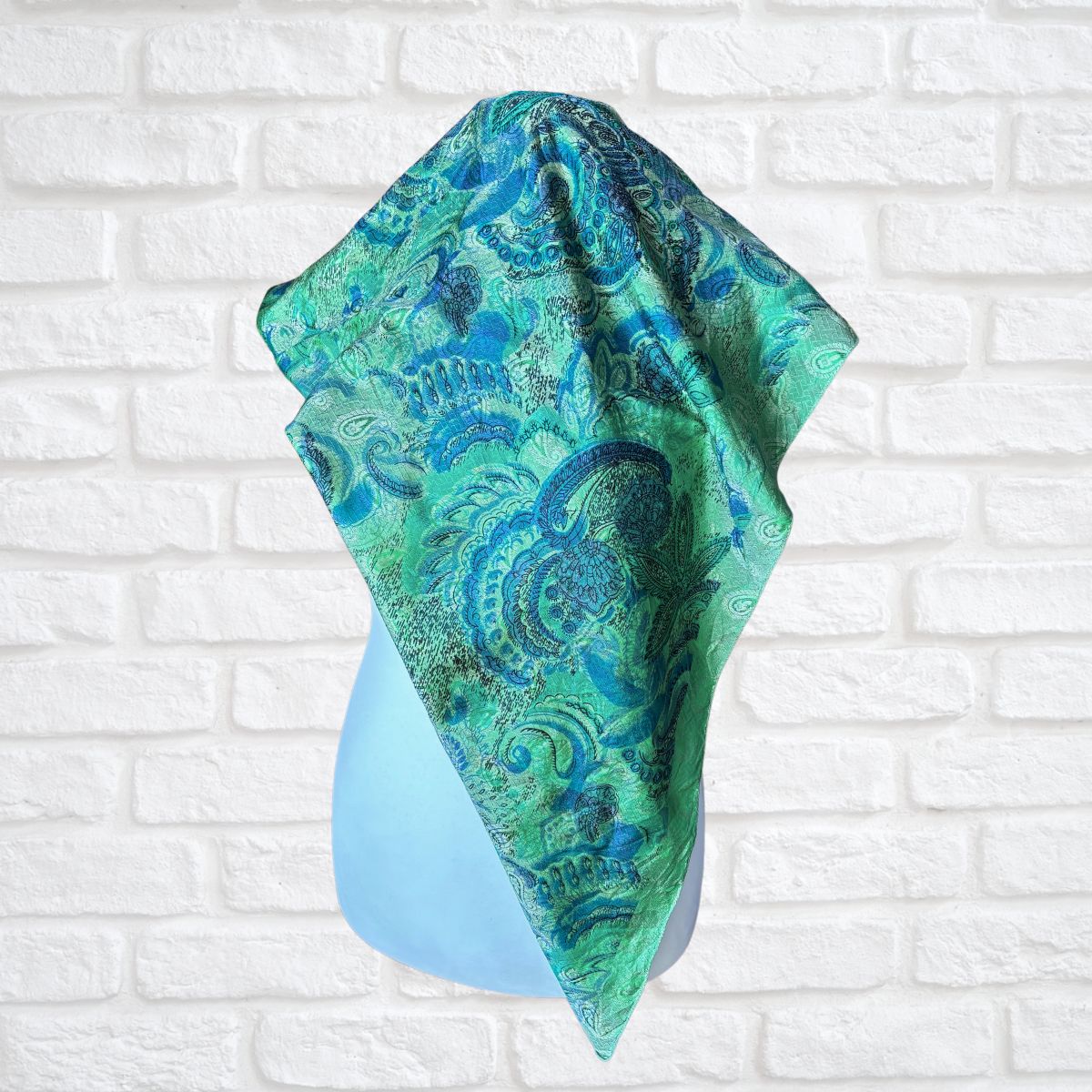 Green and blue patterned scarf  on a light blue stand against a white brick wall.