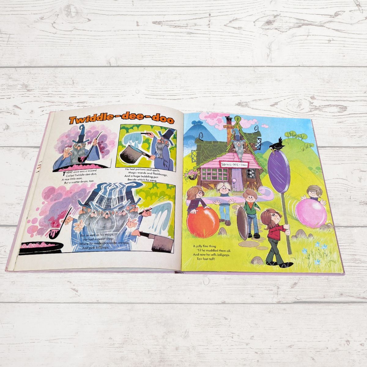 An open children's book spread laid flat on a white wood surface. The left page has colorful comic-style illustrations and text, and the right page shows a whimsical illustrated scene with children, balloons, a house, and a large purple pole.