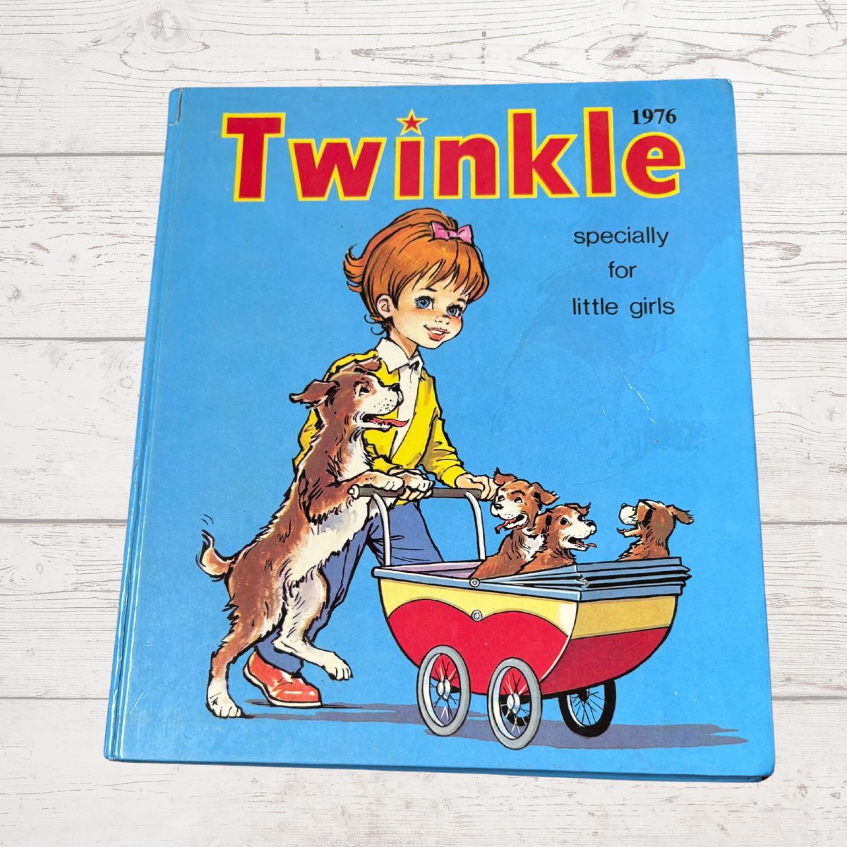 A vintage 1976 children's book titled 'Twinkle specially for little girls' featuring an illustration of a girl pushing three dogs in a red and yellow pram, placed on a white wood-plank surface.
