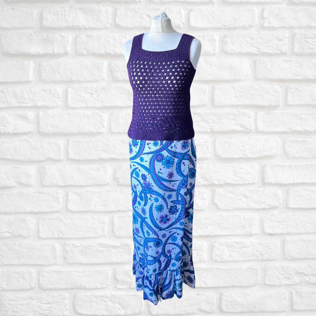 Mannequin wearing a purple sleeveless top and blue patterned skirt against a white brick wall.