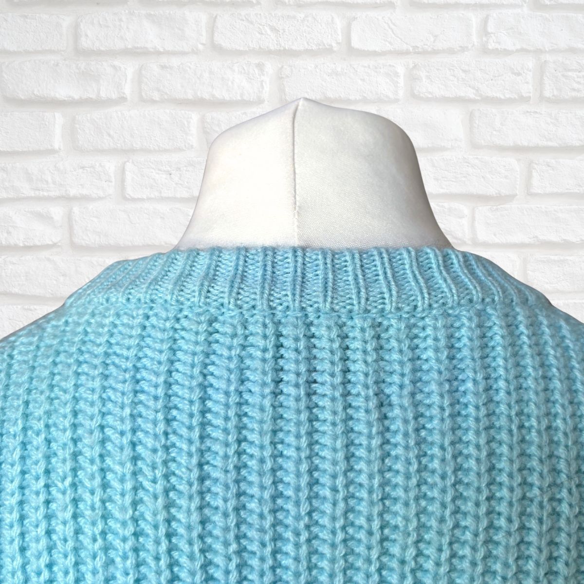 A light blue chunky knit sweater shown from the back on a white mannequin torso, set against a white painted brick wall background.