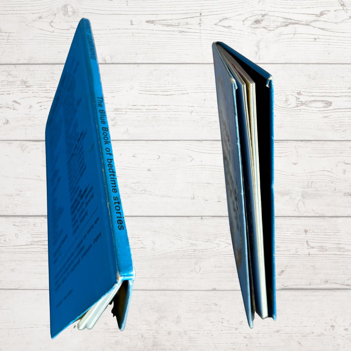 A hardcover book shown from two angles on a light gray wood plank background. One view shows the blue front cover with white title text, and the other shows the blue spine with the title 'The Blue Book of Fiddling Stories.