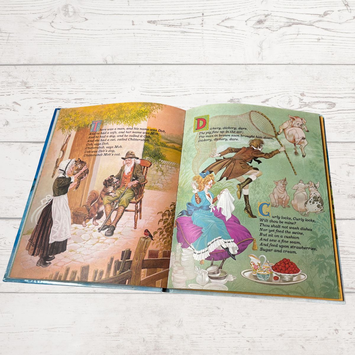 An open children's book with illustrated pages, photographed flat on a white wood surface. The left page shows a colorful historical scene with text, and the right page shows a whimsical scene with text and illustrations.